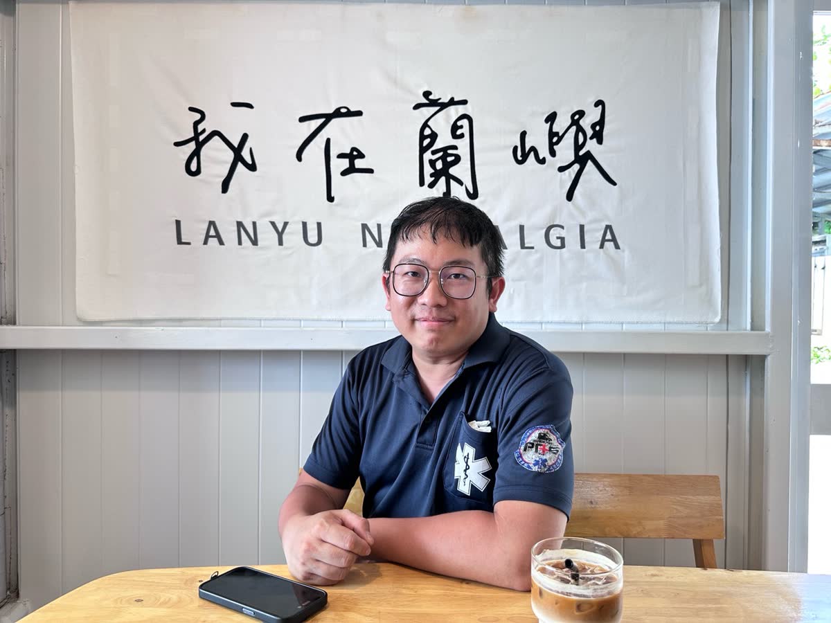 姜尚佑身穿 EMT 救護工作服，坐在蘭嶼咖啡店桌前微笑的個人照